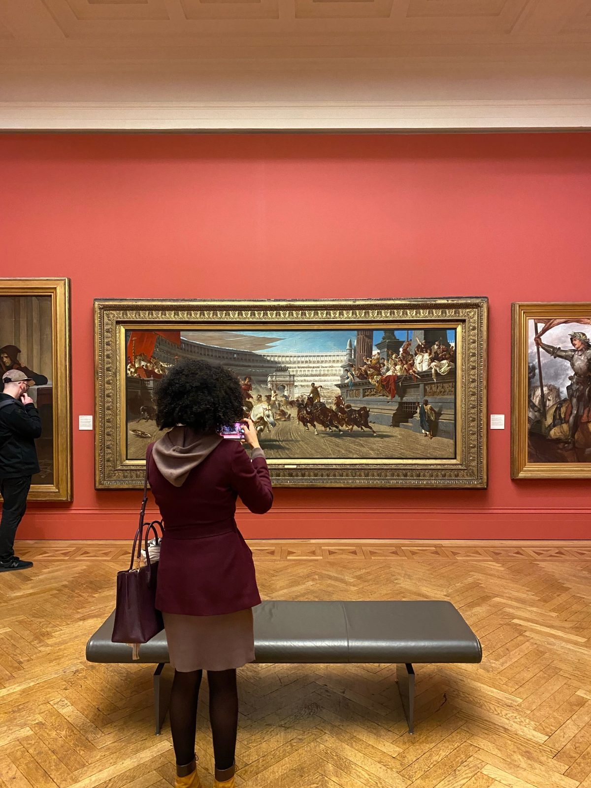 Visitor photographing a large historical painting on a red gallery wall