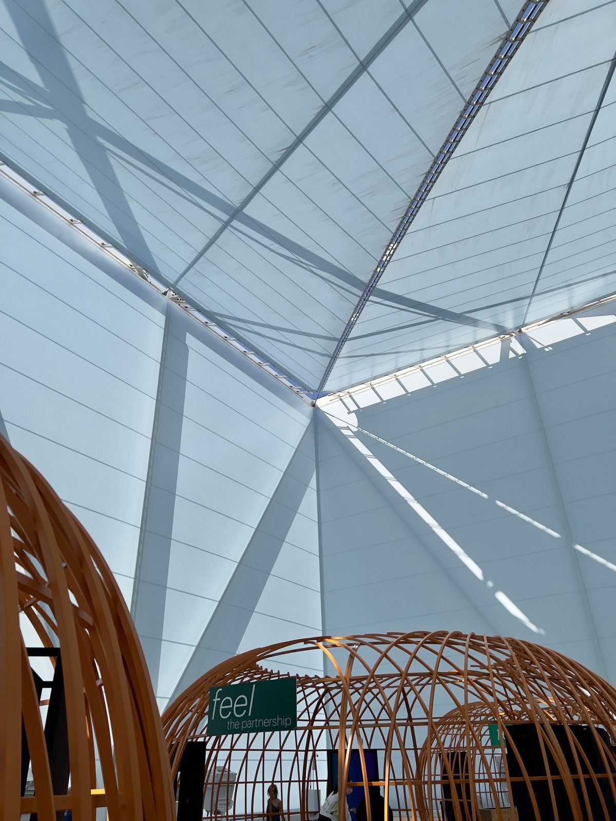 Curved timber pavilion structures beneath a white tensile roof at a design exhibition