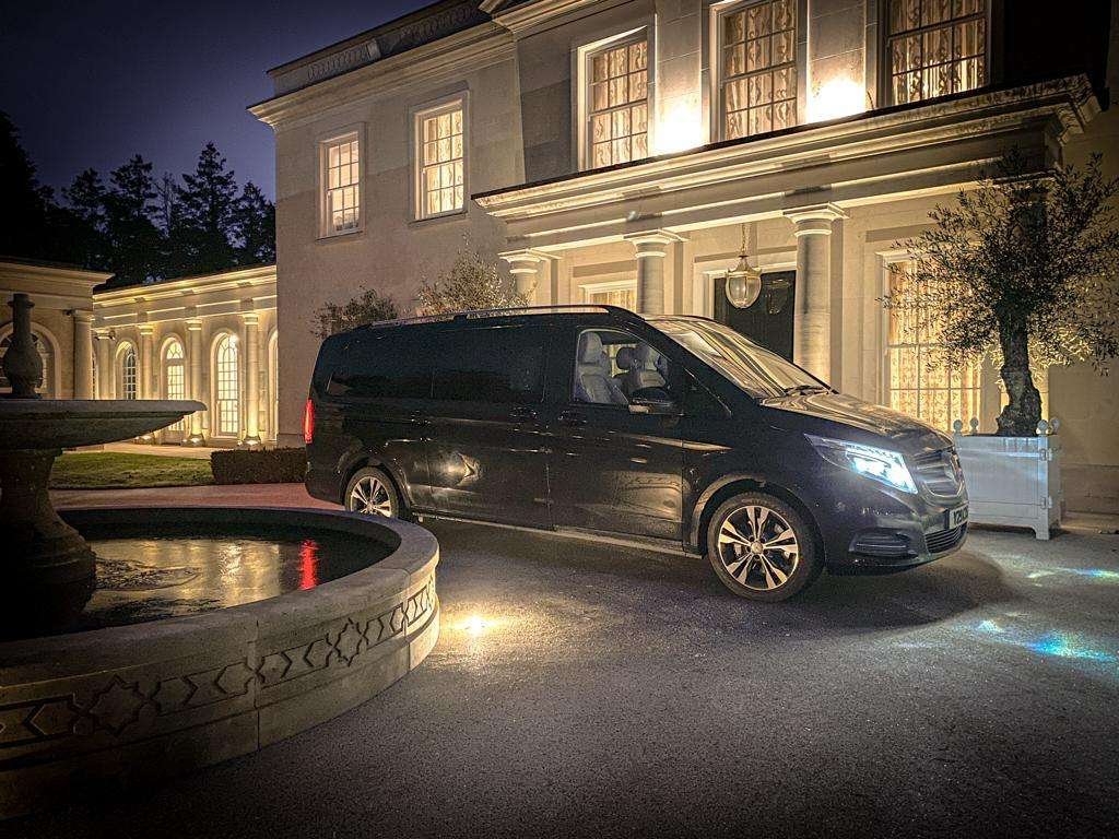 Black Mercedes-Benz V-Class family chauffeur vehicle waiting outside an illuminated luxury residence at night